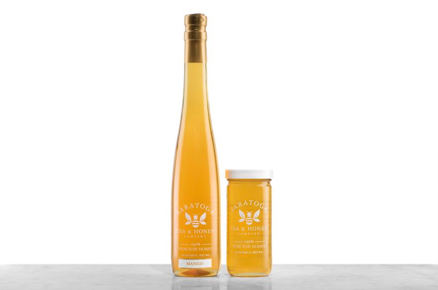 Golden Mango Infused Honey displayed in two glass jars with the logo and the words: "100% Pure Raw Honey". Left: Tall, 19oz bottle shaped like a wine bottle and capped with a cork. Right: 12oz cylindrical bottle with white cap. 