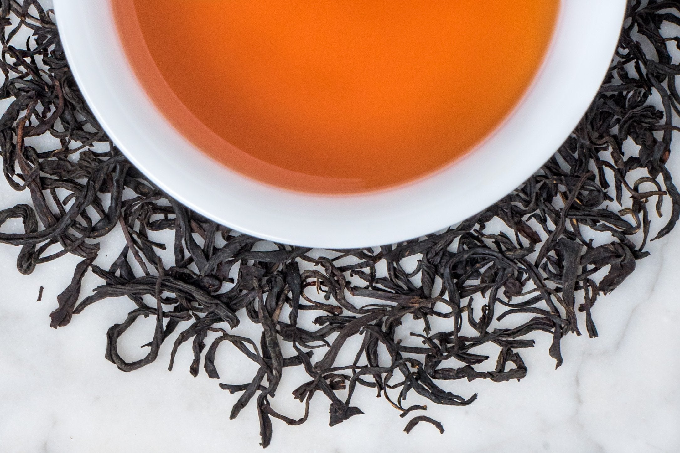 Mao Feng leaves and brewed tea. Rolled and twisted long black tea leaves surround a cup of russet-hued brewed black tea.