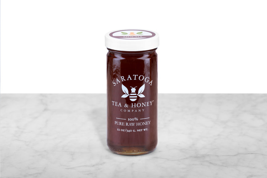 Dark brown Mocha Honey displayed in a 12oz cylindrical glass jar with a white lid. The logo and the words: "100% Pure Raw Honey" are printed on the jar.