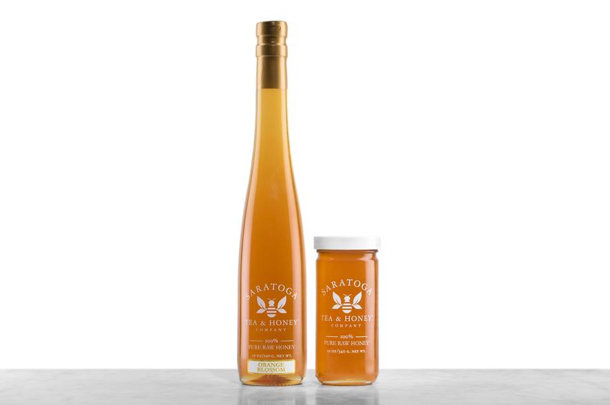 Light-amber Orange Blossom Honey displayed in two glass jars with the logo and the words: "100% Pure Raw Honey". Left: Tall, 19oz bottle shaped like a wine bottle and capped with a cork. Right: 12oz cylindrical bottle with white cap. 