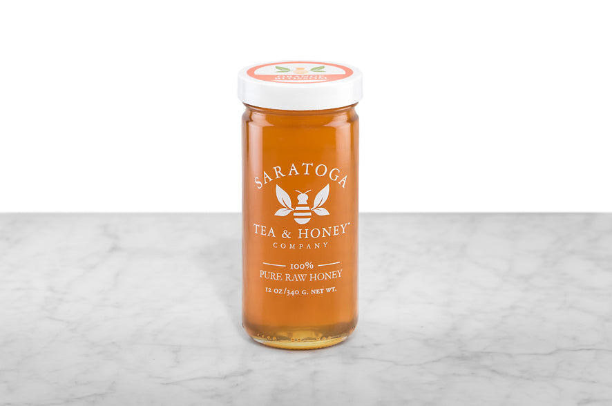 Golden amber-hued Orange Blossom Honey displayed in a 12oz cylindrical glass jar with a white lid. The logo and the words: "100% Pure Raw Honey" are printed on the jar.