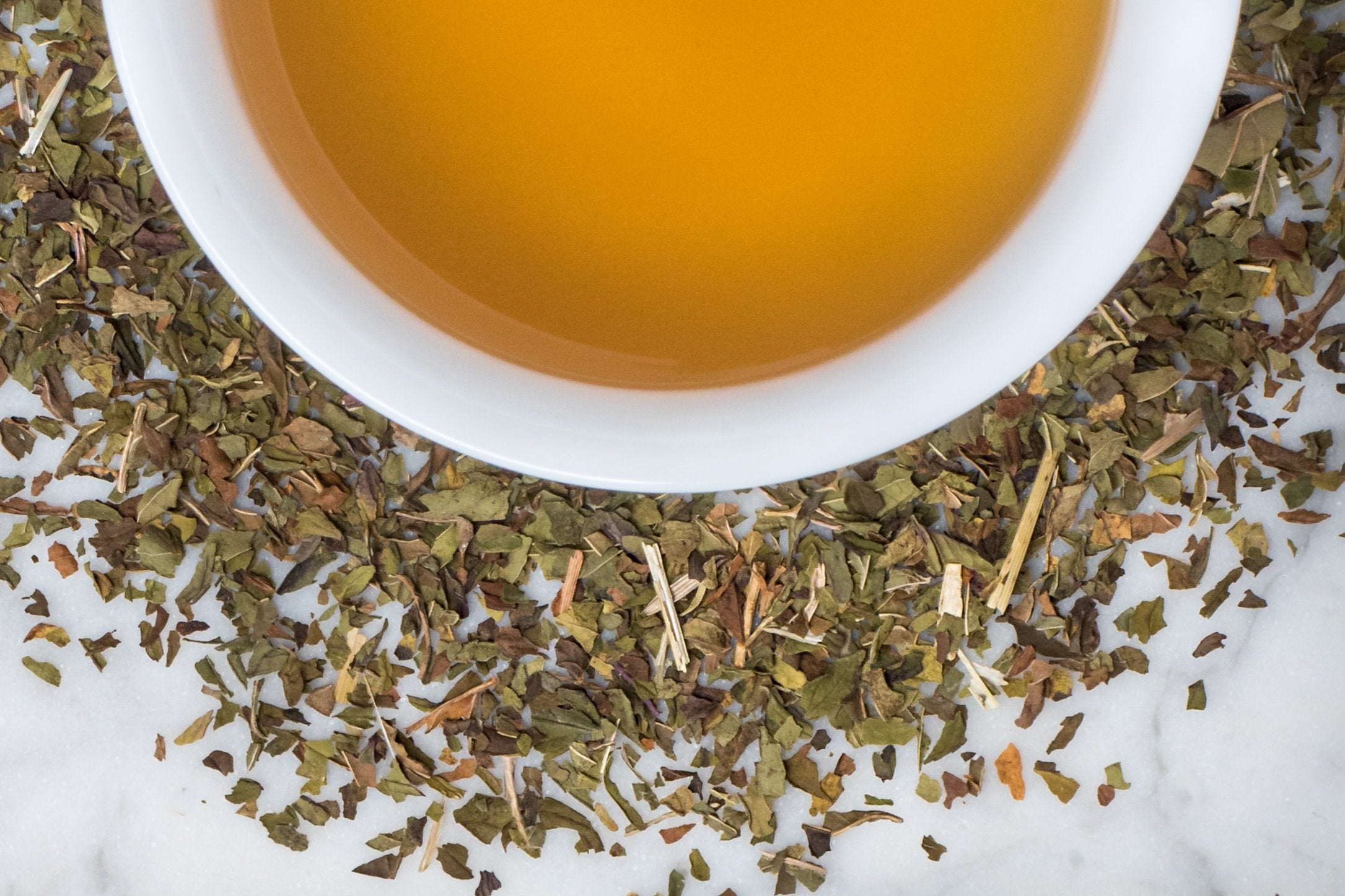 Peppermint leaves and brewed tea. Small, cut up green and brown peppermint leaves surround a cup of dark khaki-colored tea.