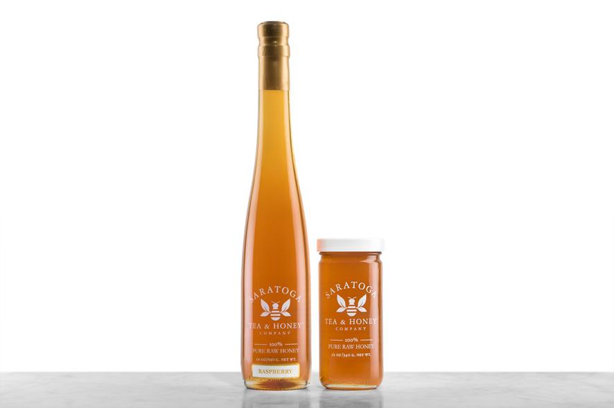 Amber-hued Raspberry Blossom Honey displayed in two glass jars with the logo and the words: "100% Pure Raw Honey". Left: Tall, 19oz bottle shaped like a wine bottle and capped with a cork. Right: 12oz cylindrical bottle with white cap. 
