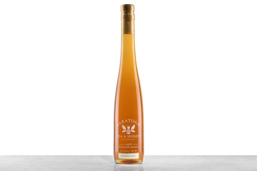 Amber-hued Raspberry Blossom Honey displayed in a tall, 19oz glass bottle shaped like a skinny wine bottle and capped with a cork. Bottle is printed with the logo and the words: "100% Pure Raw Honey".