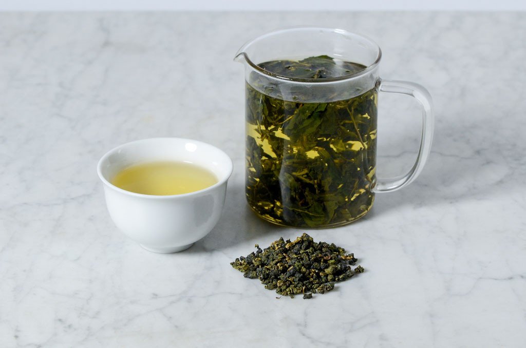 white cup of oolong next to a small pile of dry loose leaf oolong that looks like small balls and a glass infuser of brewed oolong showing how the large leaves open up during brewing