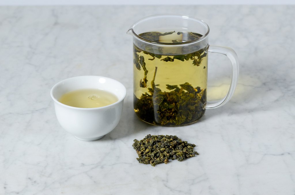 brewed Nantou Four Seasons oolong in a glass infuser and white cup alongside tightly rolled oolong leaves