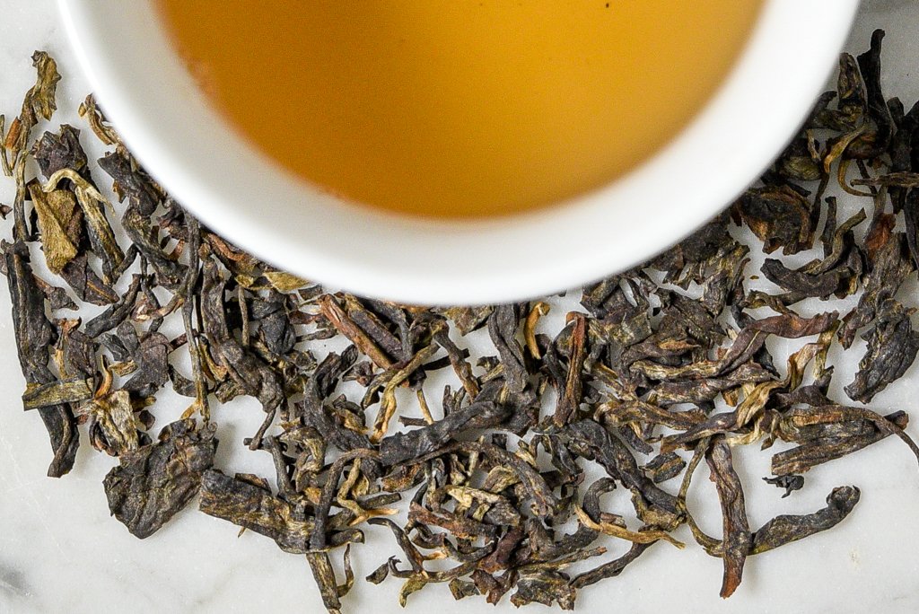 2007 Yiwu Jing Long leaves and brewed tea. Green, brown, and gold flattened and twisted leaves surround a cup of brewed amber-gold aged tea.
