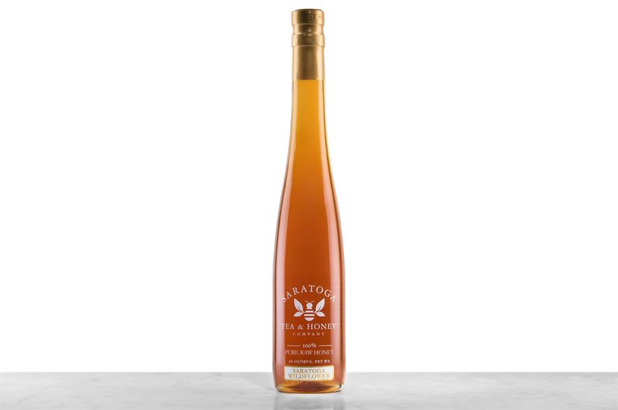 Amber-hued Saratoga Springs Wildflower Honey displayed in a tall, 19oz glass bottle shaped like a skinny wine bottle and capped with a cork. Bottle is printed with the logo and the words: "100% Pure Raw Honey".