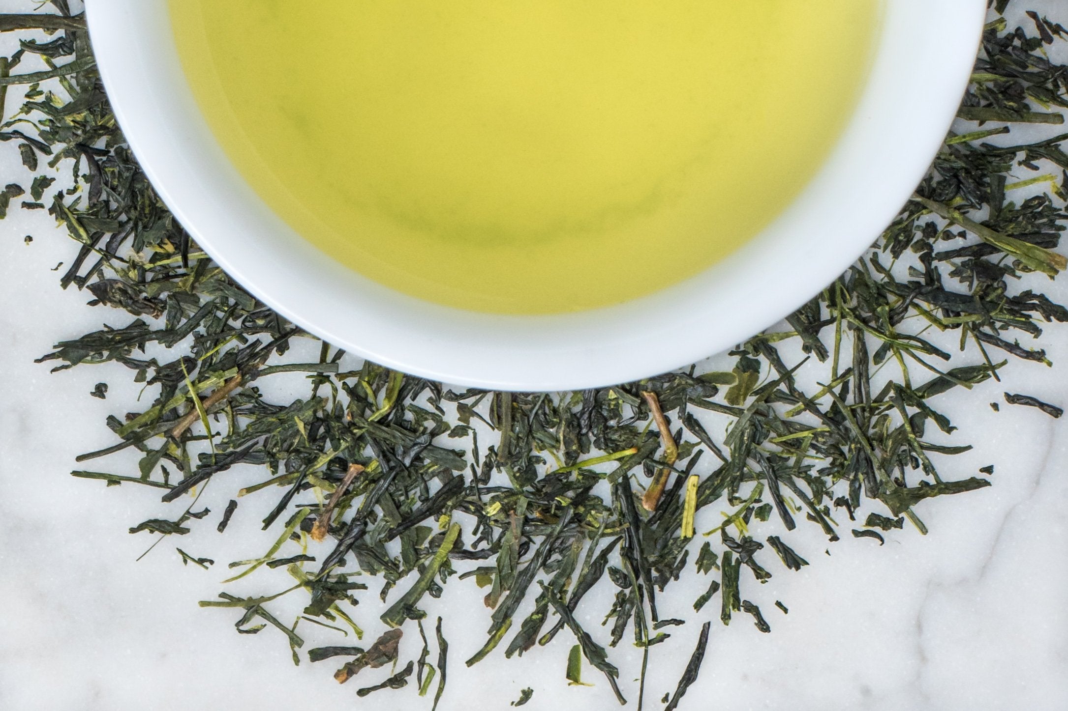 Sencha Sumire leaves and brewed tea. Thin and short dark green leaves surround a cup of brewed bright, yellow-green tea.
