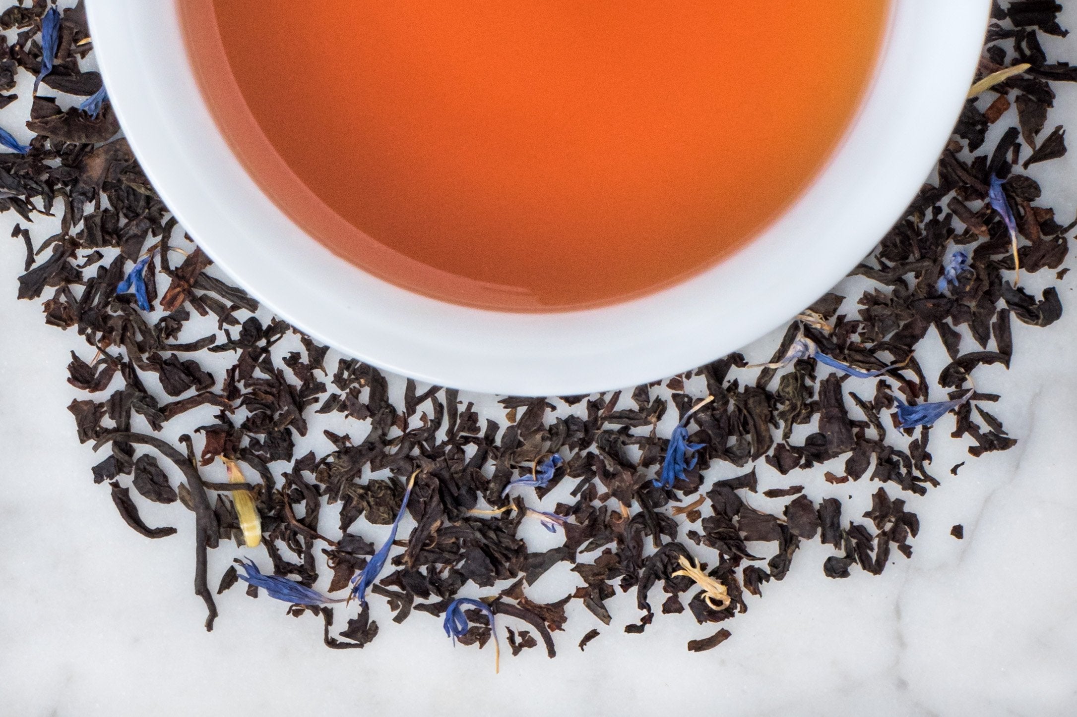 Spa City Earl leaves and brewed tea. Small black tea leaves and blue cornflower petals surround a cup of russet-brown brewed black tea.