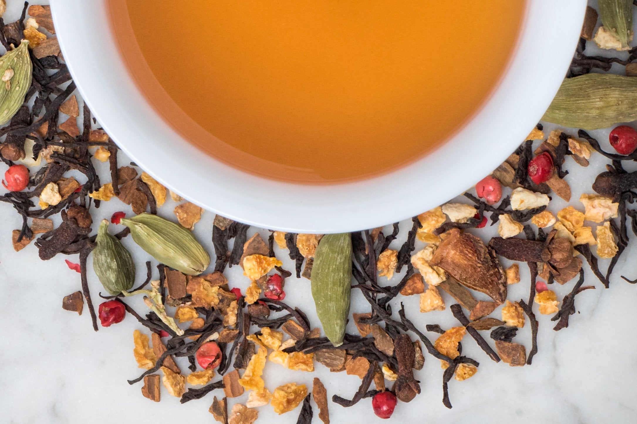 Spiced Orange Chai leaves and brewed tea. Thin, twisted black tea leaves and spices surround a cup of golden-amber hued chai tea.