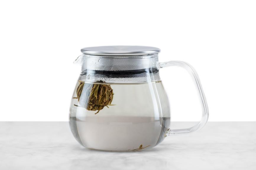 Jasmine Flower Craft tea ball brewing stage 2 - steeping. Ball of green tea floats in clear glass teapot with a few leaves starting to pull away from the ball. 