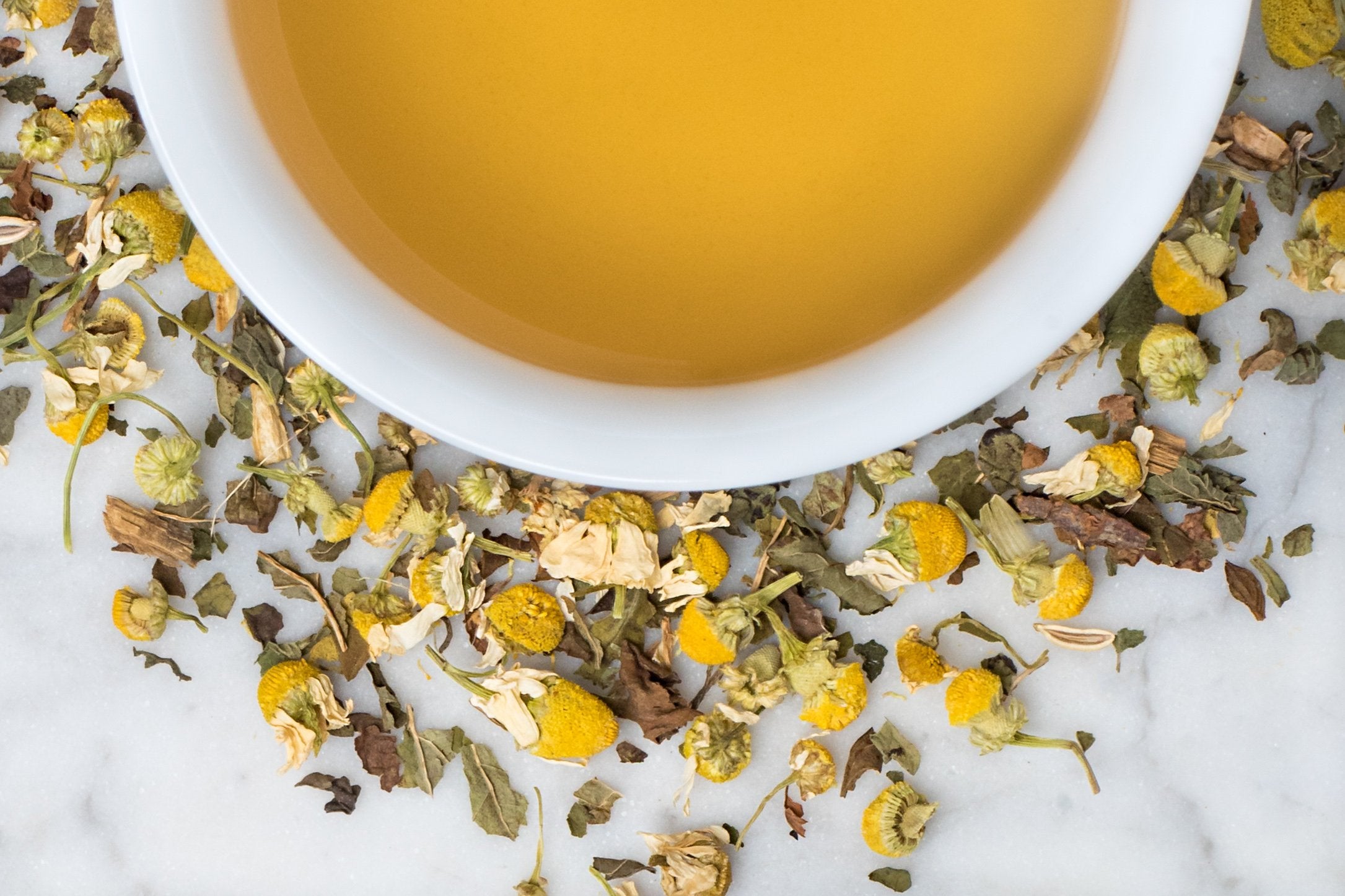 Tummy Tonic leaves and brewed tea. Leaves and pillowy camomile buds surround a cup of old-gold hued brewed herbal tea.