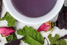 rare purple tea infusion displayed in a white cup surrounded by whole rosebuds, basil, and raspberry leaf ingredients