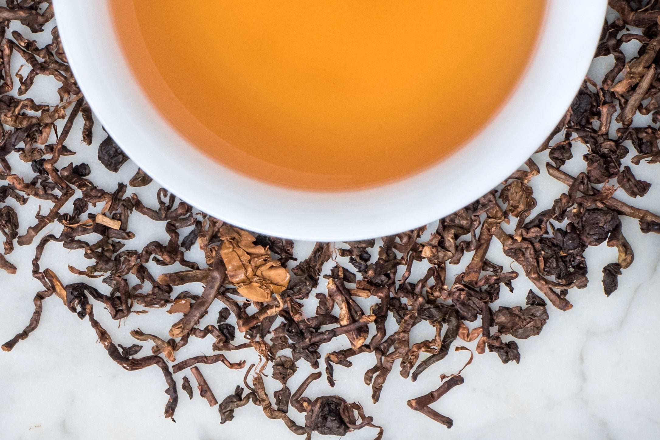 Wood Dragon leaves and brewed tea. Twigs of brown stem withs a few brown leaves surround a cup of amber-colored oolong tea.
