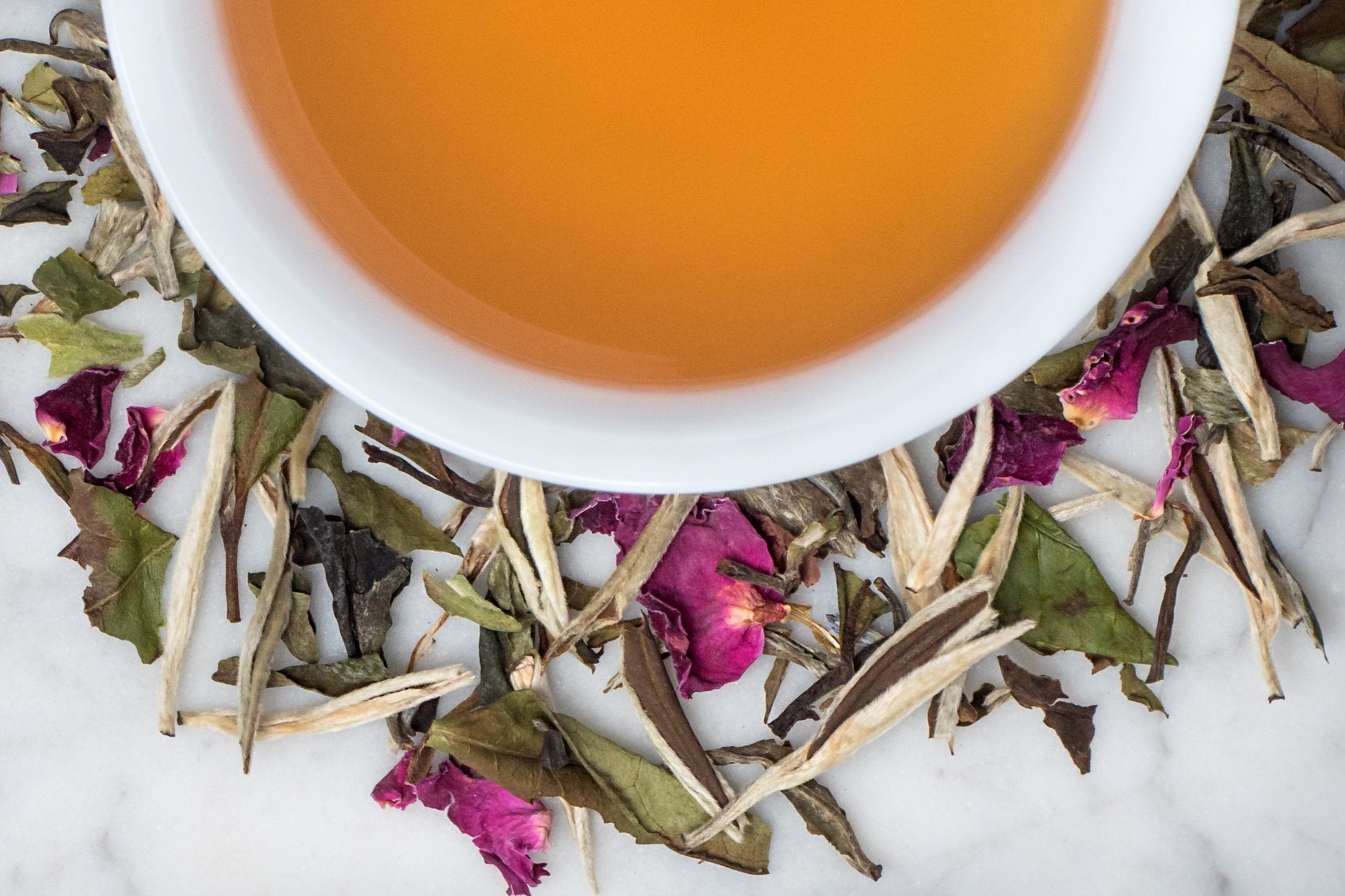 Yaddo Rose Garden leaves and brewed tea. Long downy tea buds, torn leaves, and pink rose petals surround a cup of light amber-hued brewed tea.