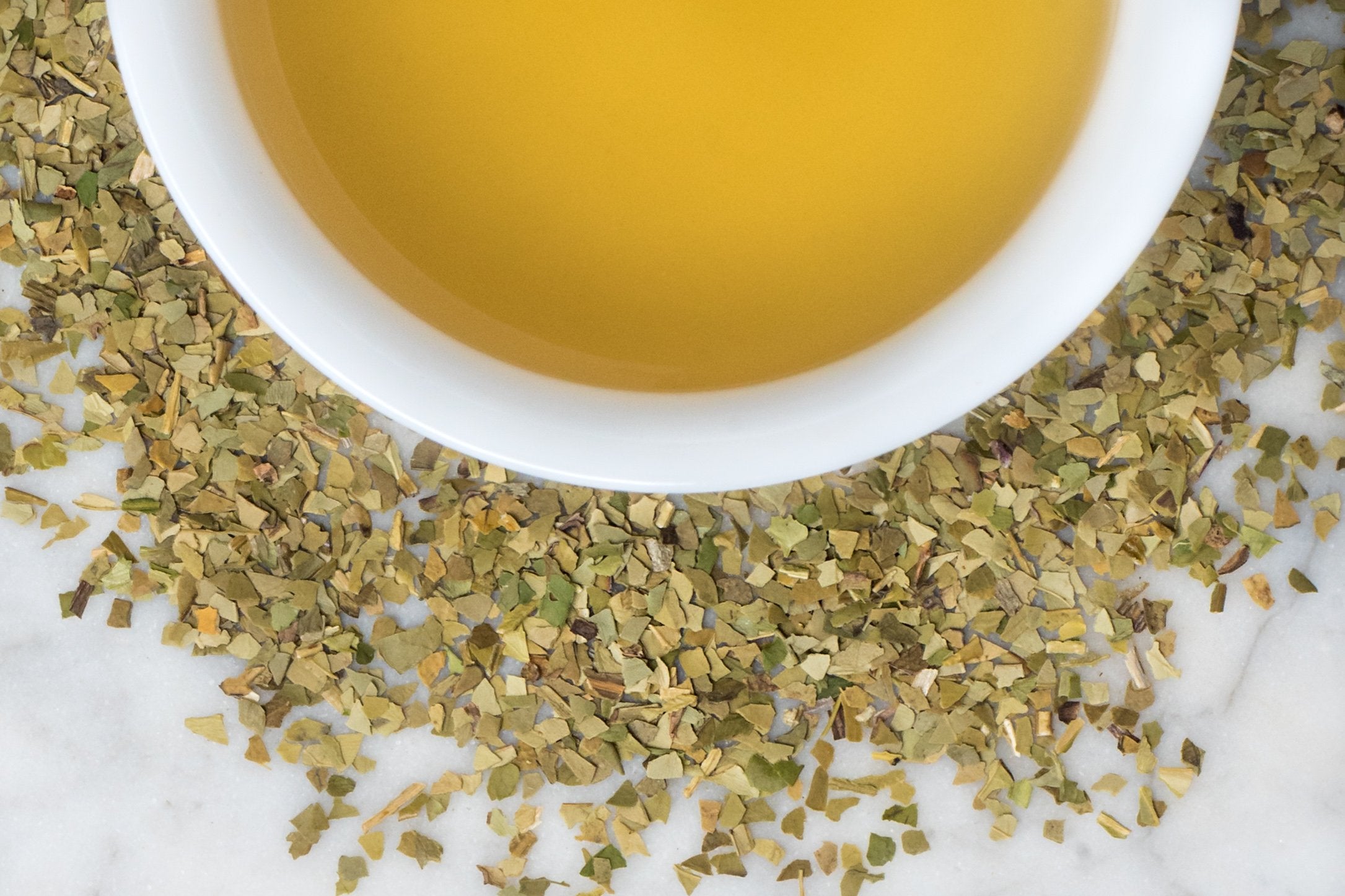 Yerba Mate leaves and brewed tea. Small, confetti-like green and khaki Yerba Mate leaves surround a cup of brownish-yellow brewed tea. 