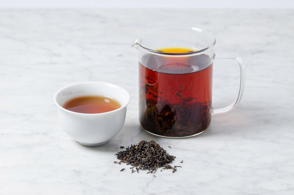 rich red english breakfast black tea infusion shown in a white cup next to a glass infuser and a pile of loose leaf black tea