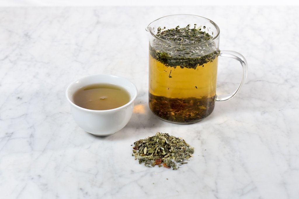 Baby bump pregnancy tea brewed and presented in a white cup next to a glass infuser with a pile of loose herbal tea
