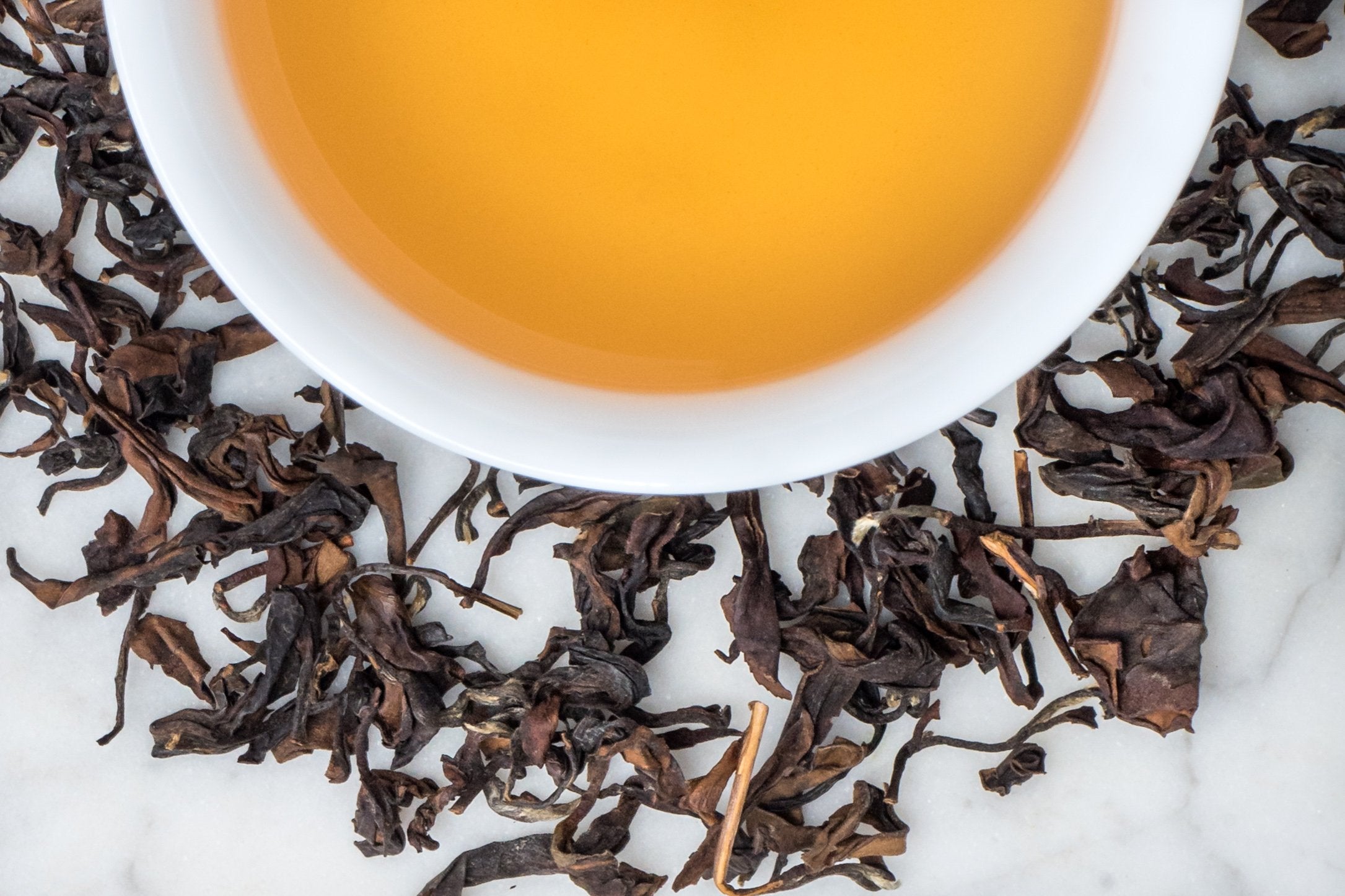 Bai Hao Jingmai leaves and brewed tea. Long, twisted leaves and stems of various dark and medium-brown hues surround a cup of yellow-gold colored brewed oolong tea.
