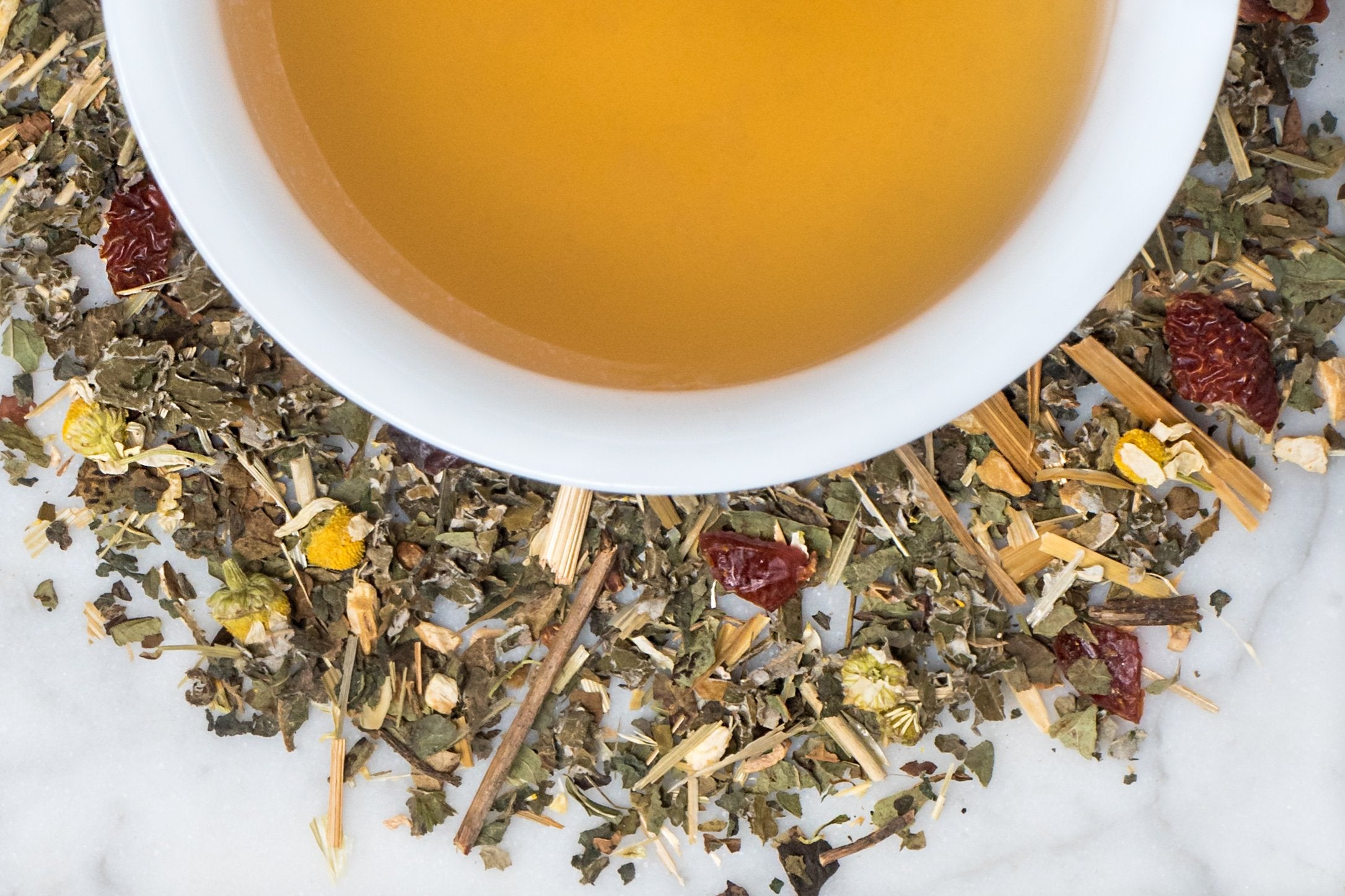 Bonjour Bébé leaves and brewed tea. Nettle, oat straw, and red raspberry leaf tea surrounds a cup of light golden-brown brewed tea.