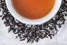 Coco Shou leaves and brewed tea. Long, twisted black tea leaves and tiny chocolate kisses surround a cup of dark-brown brewed tea.