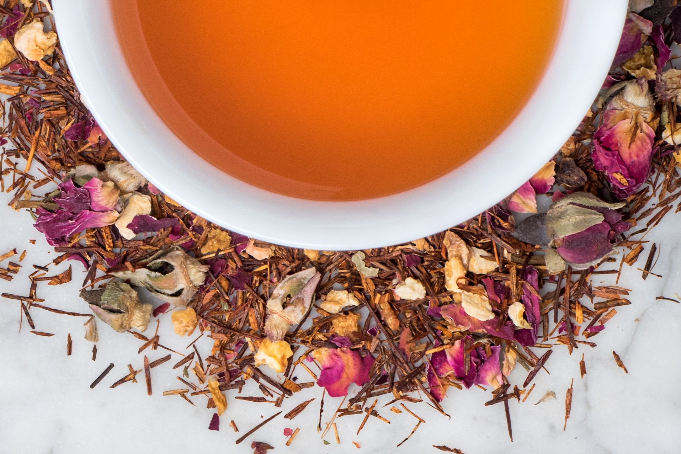 Egyptian Goddess leaves and brewed tea. Rooibos, rose and orange peel surround a cup of brewed reddish-brown herbal tea.