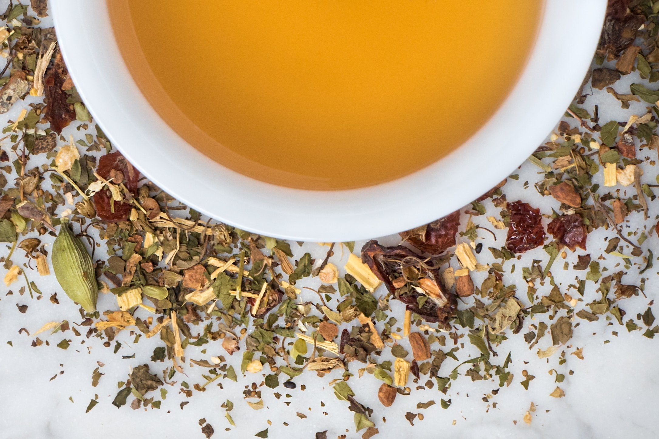 Exhale leaves and brewed tea. Small leaves, berries, and cardamom pods surround a cup of amber-hued brewed herbal tea.