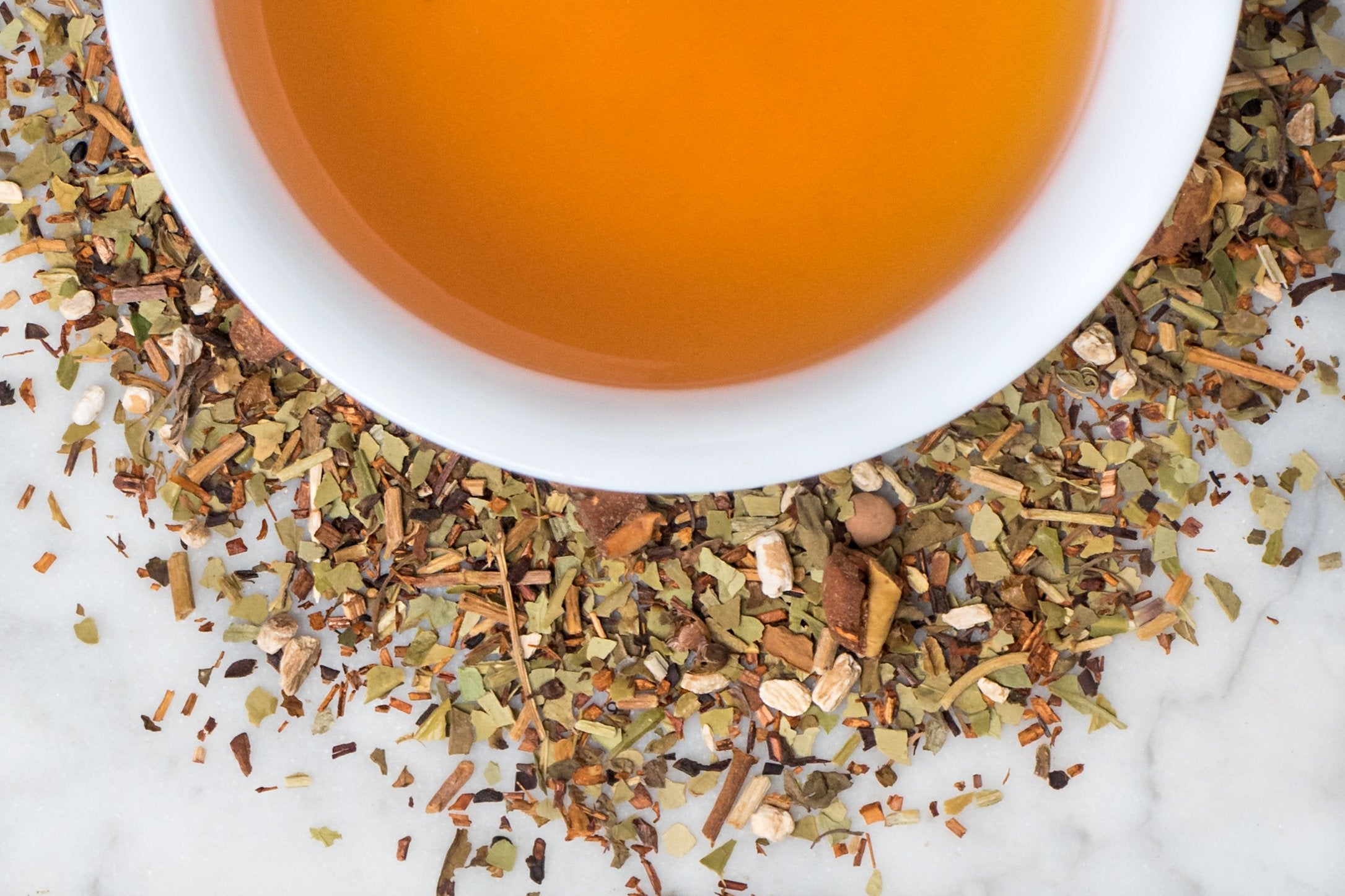 Focus Pocus leaves and brewed tea. Small, khaki green yerba mate leaves and other leaves and spices surround a cup of amber-hued brewed tea.