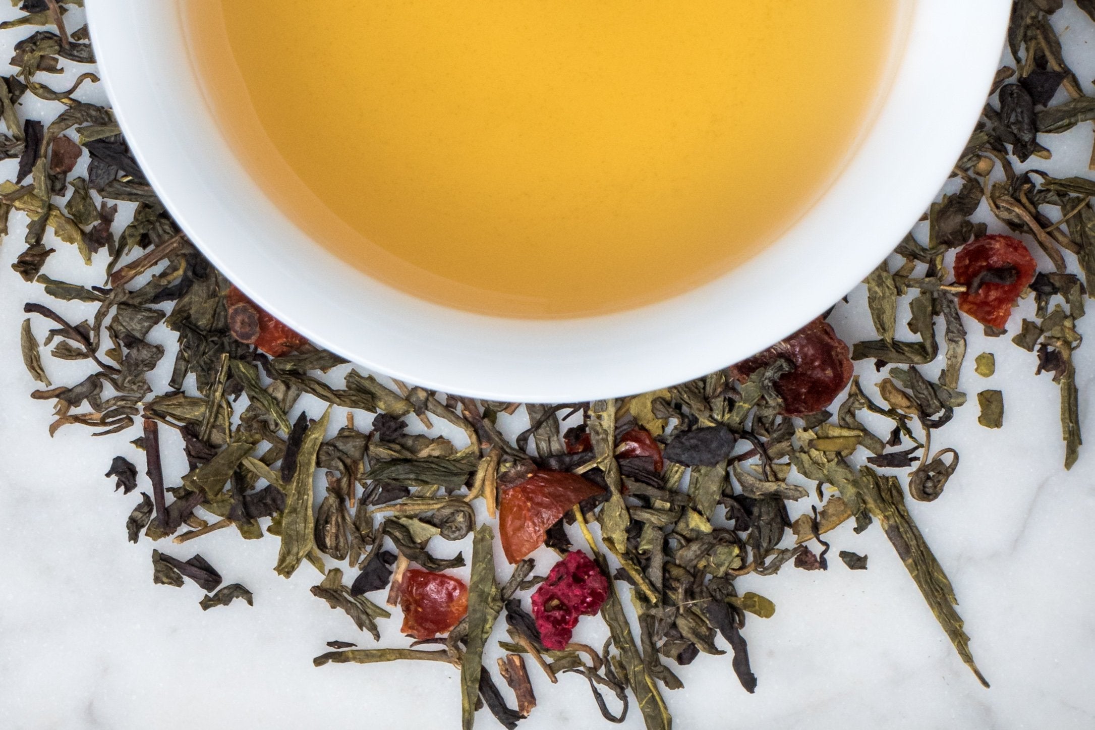 Green Paddock Pom Pom leaves and brewed tea. Black and green tea leaves and dried fruit surround a cup of light gold-colored brewed tea.