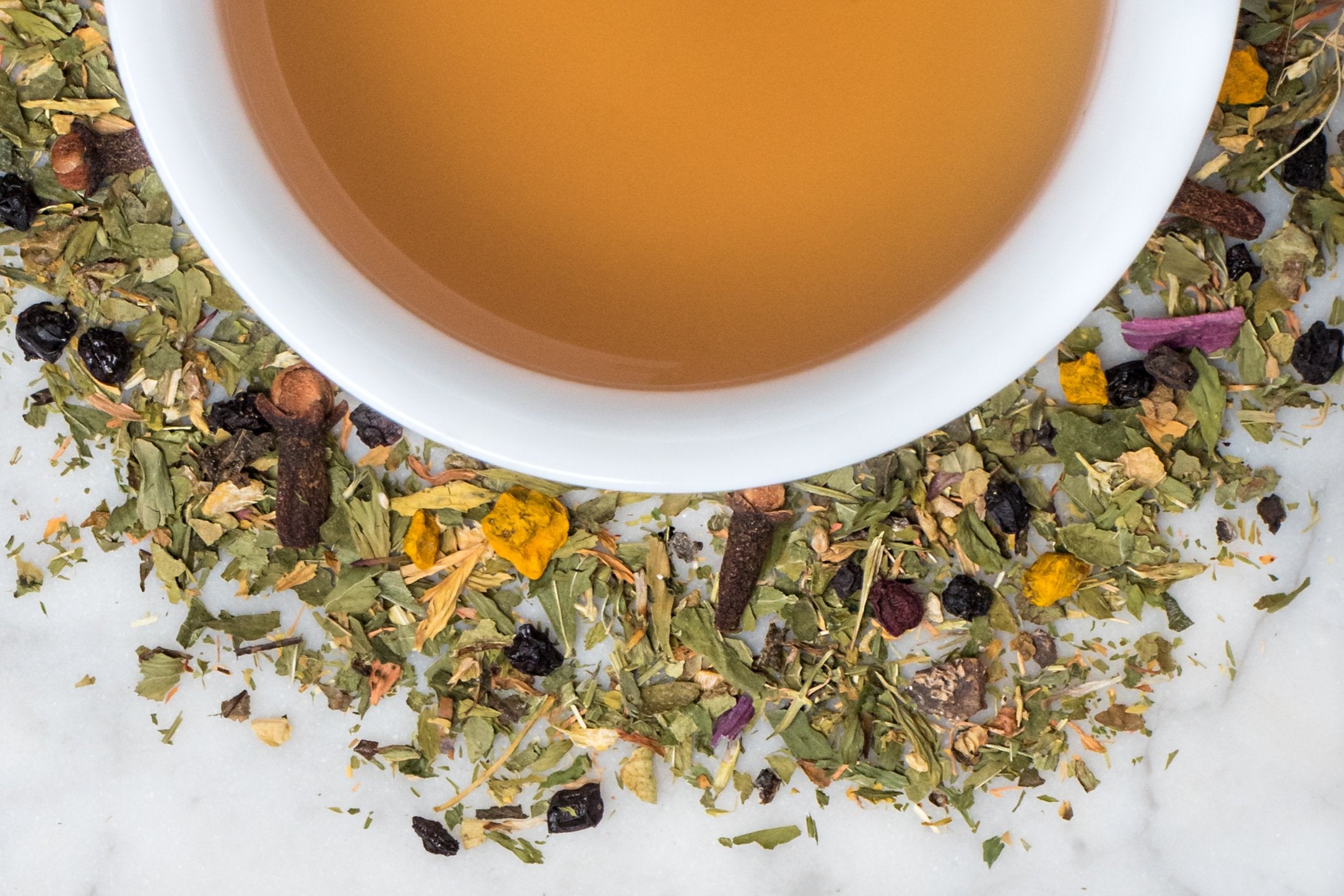 Health Potion herbal tisane leaves and brewed tea. Torn, green leaves, pieces of turmeric and ginger, and dried berries surround a cup of brewed cognac-colored tea.