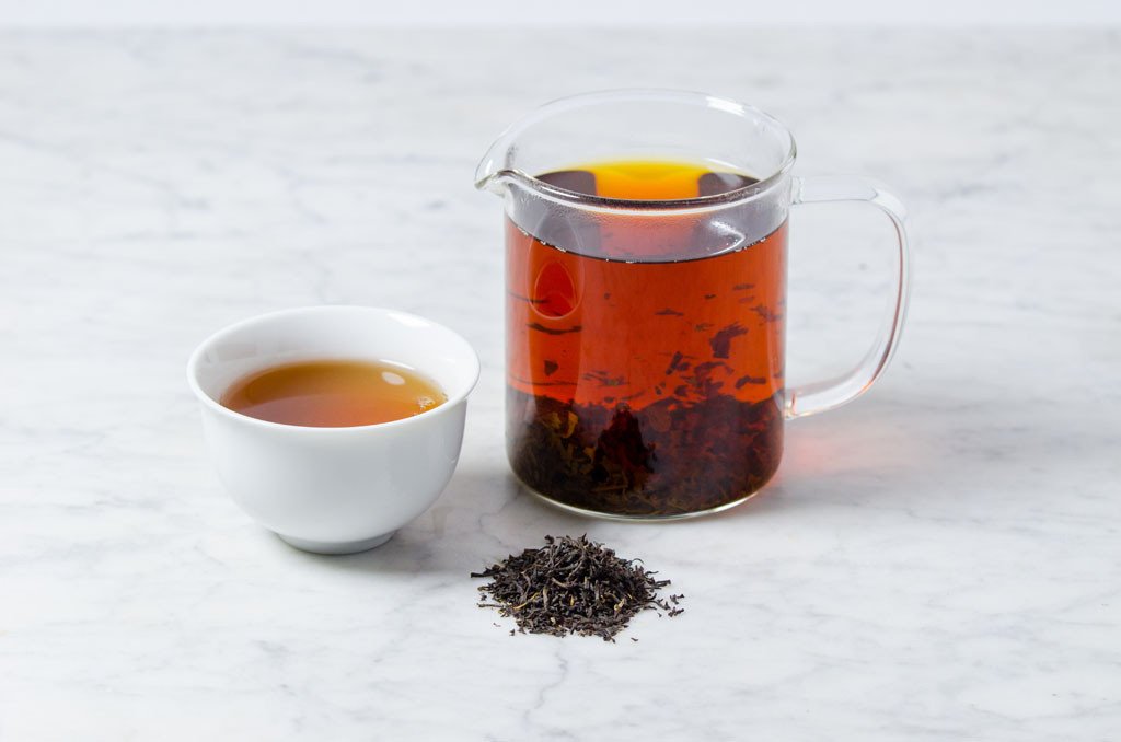 Rich amber Kenya Kangaita brew in a white cup next to a glass infuser and pile of loose leaf black tea leaves
