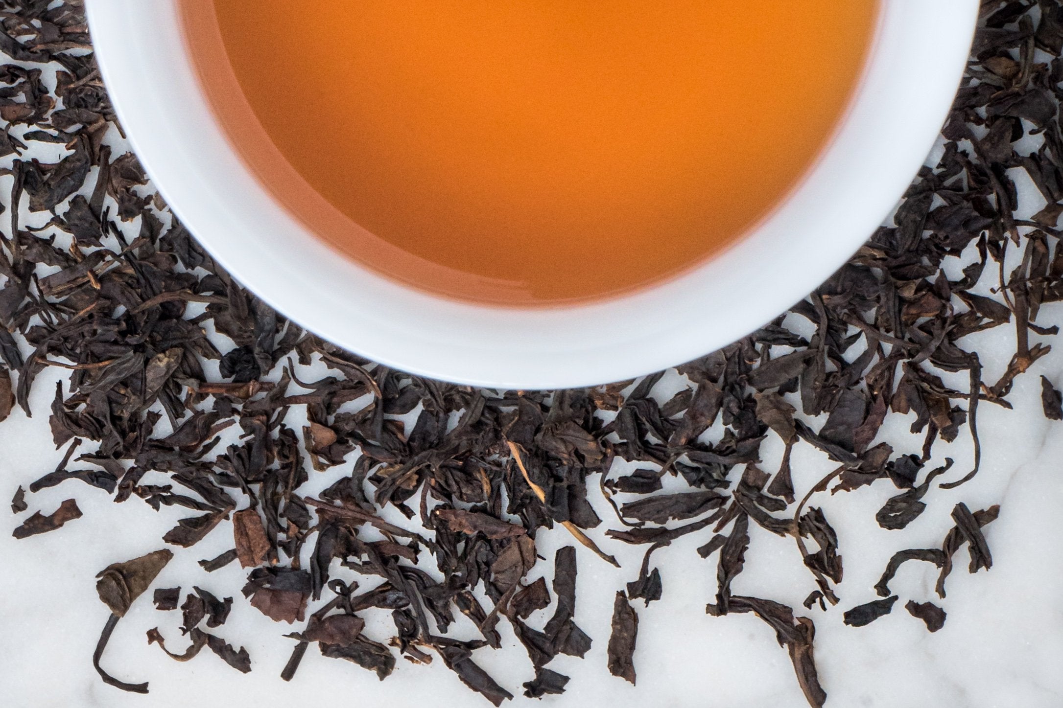 Lapsang Souchong leaves and brewed tea. Small, twisted smoked black tea leaves surround a cup of dark-amber brewed tea.