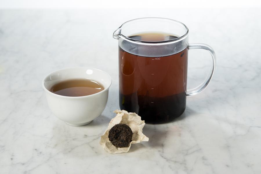 unwrapped mini pu'er tea cake (toucha) next to a white cup and glass infuser filled with 2012 Menghai aged tea
