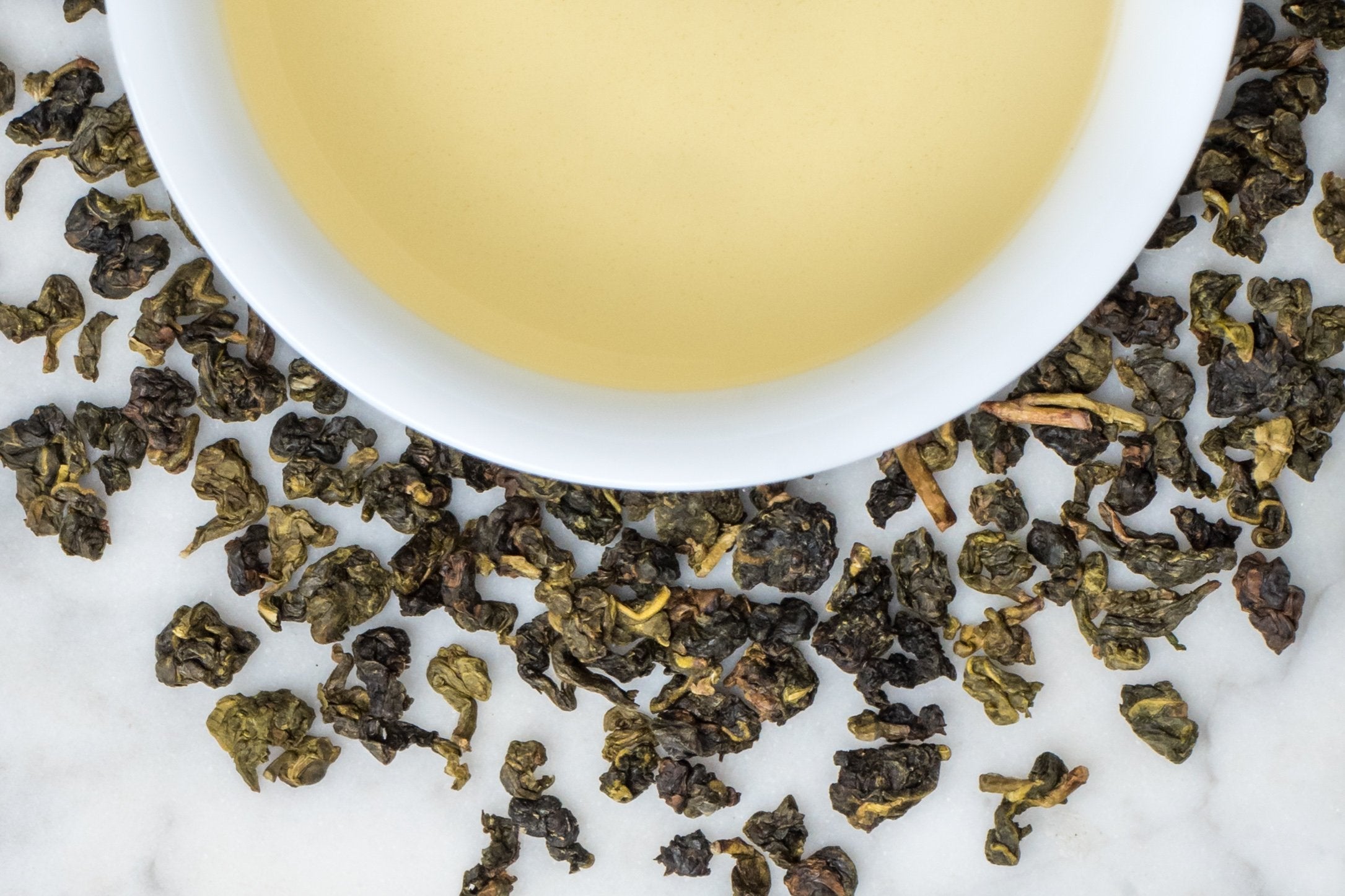 Nantou Four Seasons leaves and brewed tea. Small, tightly rolled balls of light and dark green oolong surround a cup of very pale yellow brewed tea.