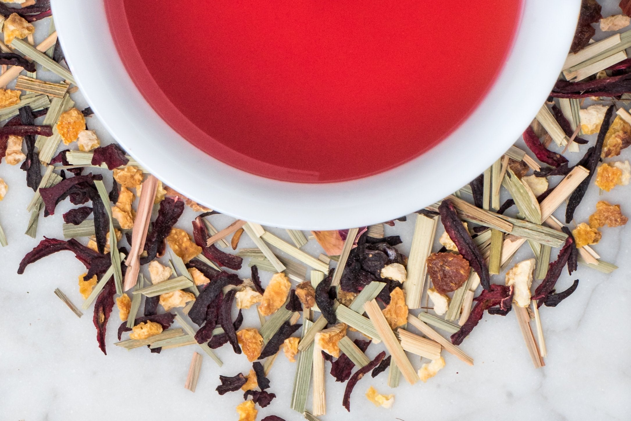 Saratoga Sunrise leaves and brewed tea. A mix of lemongrass, citrus peel, and hibiscus surrounds a cup of vibrantly pink brewed tea.