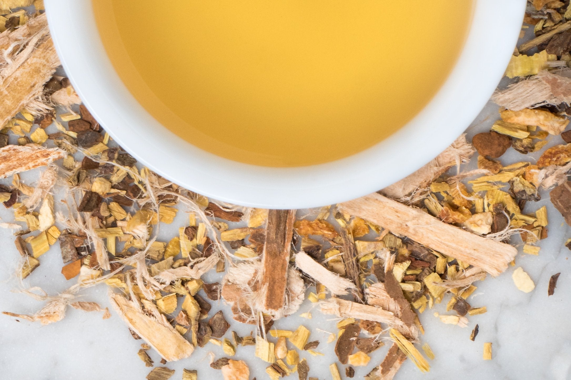Songbird leaves and brewed tea. Fuzzy strips of slippery elm bark, cinnamon, orange peel, ginger, and licorice surround a cup of golden-hued brewed throat coat tea.