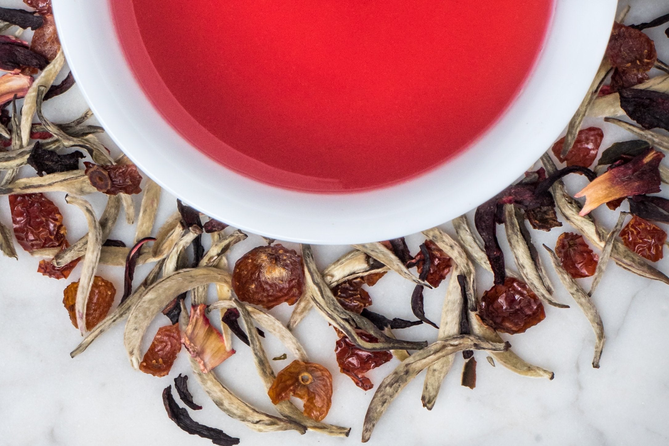 The Canfield in Red leaves and brewed tea. Downy white tea buds, rosehips, and hibiscus flowers surround a cup of deep pink brewed tea.