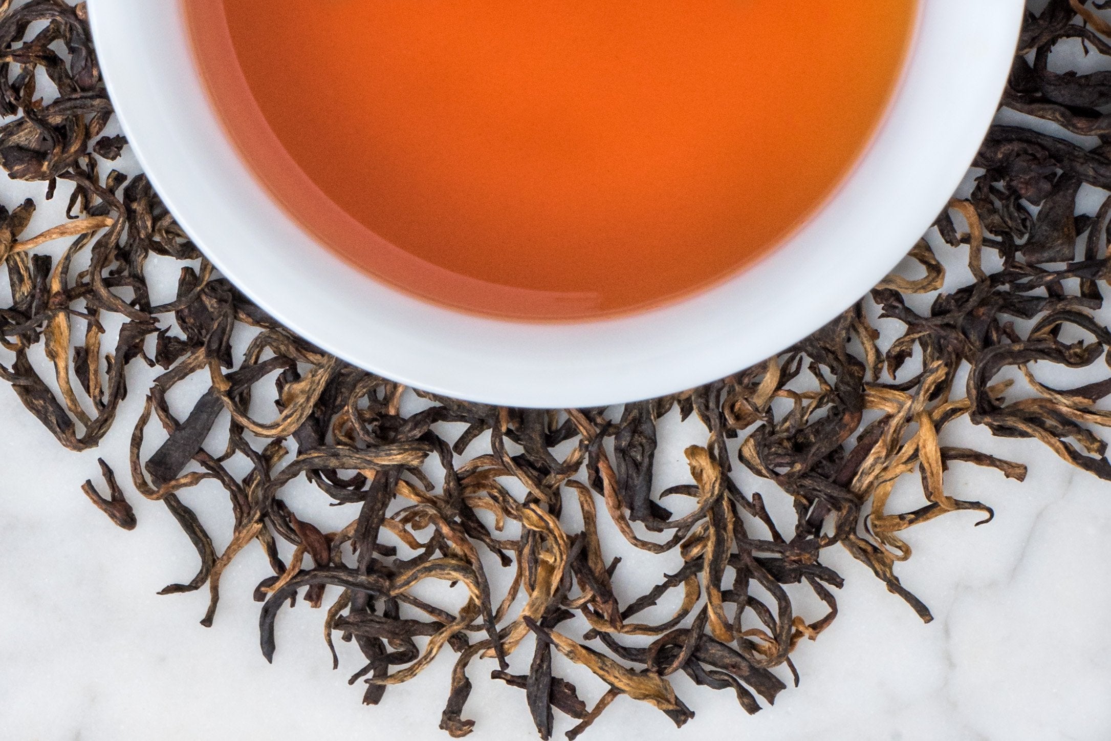 Yunnan Da Ye leaves and brewed tea. Twisted dark brown leaves and golden brown buds surround a cup of reddish-brown brewed black tea.